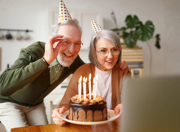 Beautiful senior couple in party hats celebrating birthday at home online on laptop, receiving congratulations from family trough video chat, happy man waving at camera, woman holding chocolate cake