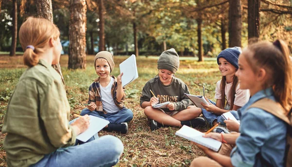 Group of excited school children boys and girls with young female teacher sitting on ground in forest during ecology lesson outdoors, writing down in notebooks their discoveries of exploring nature