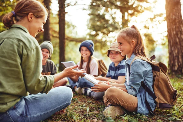 School activities in nature. Group of curious children and young female teacher sitting together on green grass in forest and learning about environment, boys and girls during ecology lesson in wood