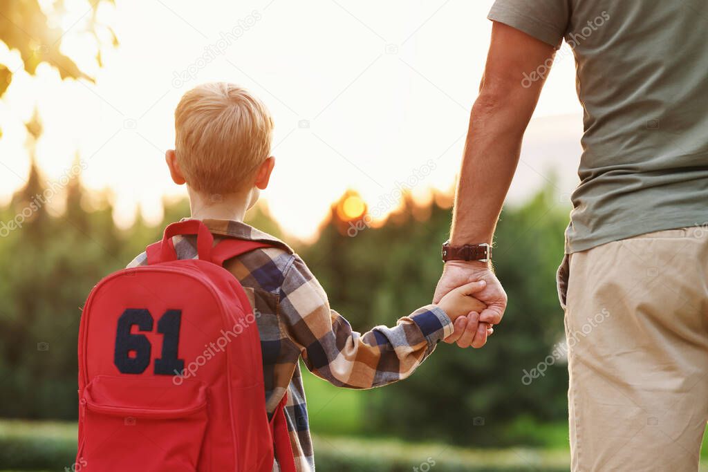 Rear view of little son schoolboy with backpack holding hand of father dad while going to first grade on sunny autumn day, cropped shot of parent and small child on the way to school, standing in park