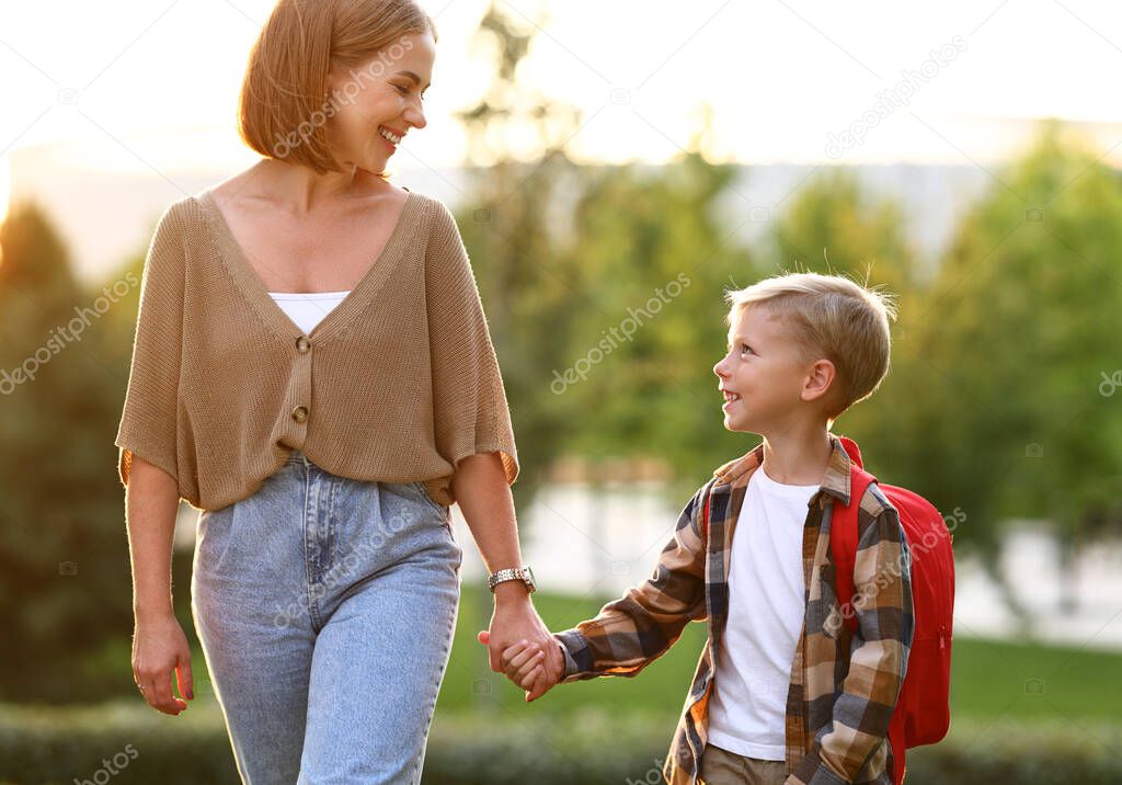 Mother and little son schoolboy with backpack holding hands, going to school together through park on sunny autumn day, cute happy boy looking at mom with smile and being ready to study