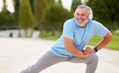 Açık hava antrenmanı sırasında mutlu, olgun sporcu, kulaklık takan, spor kıyafeti giyen, kaslarını ısıtan, tek bacağının üzerine çömelmiş, parkta aktif yaşam tarzının tadını çıkaran.