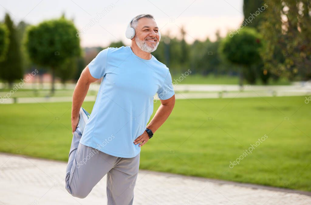 Photo of mature active positive man in wireless headphones doing legs stretching exercises with smile, warming up muscles before routine workout and jogging outside in city park in sunny morning