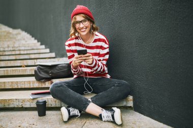 Kulaklıklı mutlu öğrenci kız elinde akıllı telefonuyla oturmuş dersten sonra kahve molası veriyor dişlek gülüşlü kameraya bakıyor çizgili komik kıyafetler ve gözlük takıyor.