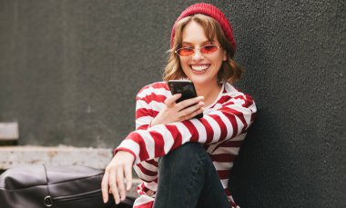 Güneş gözlüklü, elinde cep telefonuyla dışarıda sırt çantasıyla oturup komik videolar izleyen neşeli bir kız öğrenci.