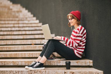 Moda kıyafetleri ve kulaklık takan çekici kız öğrencinin yan görüntüsü merdivenlerde dizüstü bilgisayarla, defterlerle oturuyor ve internetten bilgisayarına gülümseyen bir şekilde bakarken kahve fincanı alıyor.