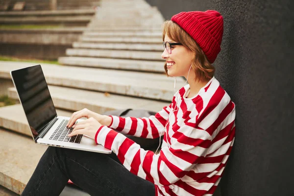 积极可爱的女学生 戴着耳机 坐在外面拿着笔记本电脑 笑着看着空荡荡的模拟屏幕 键盘上打字 和朋友们在网上聊天 穿着休闲时髦的衣服 — 图库照片