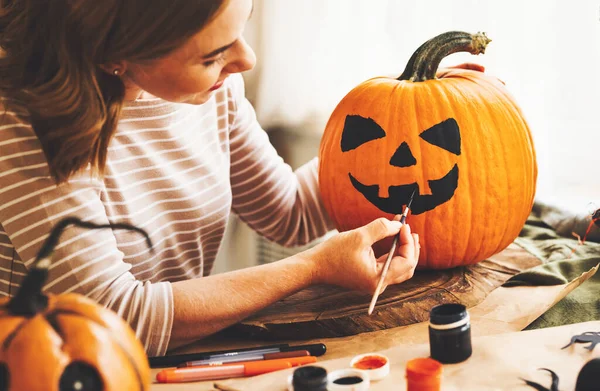 Young woman drawing scary face on orange pumpkin with paint brush for house decoration during Halloween holiday, female creating jack-o-lantern while sitting at table in kitchen at home