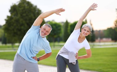 Sağlıklı zihin ve vücut. Güneşli bir sabahta parkta birlikte ısınırken spor giyim kuşamında gülümseyen olgun aile babası ve kadın kollarını esnetiyorlar. Etkin yaşam biçimi