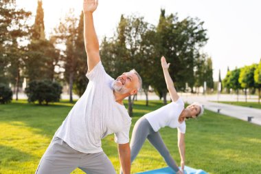 Güneş doğarken yeşil park çimlerinin üzerinde, Utthita Trikonasana üçgeninde duran pozitif emeklilerin eş-koca yogası yaparken çekilmiş yakın plan fotoğrafı ışıltılı bir gülümsemeyle yukarı bakıyor.