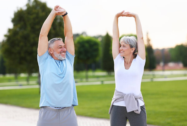 Morning exercises. Happy mature married couple, man and woman in sportswear warming up together doing stretching exercises raised hands up outdoors on park lane in morning. Healthy lifestyle concept