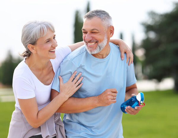 Portrait of lovely happy elderly couple on morning run outside in city park, retirees wife and husband rejoice in active lifestyle, smiling woman tenderly embracing her spouse after routine jogging