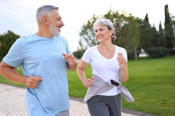 快乐的老夫妇穿着运动服在公园里跑来跑去 可爱的退休夫妇在阳光灿烂的早晨慢跑 面带微笑地互相看着对方 健康生活方式概念 — 图库照片