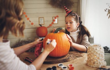 Sevimli küçük bir kız Cadılar Bayramı süslemesi yapıyor annesiyle mutlu bir aile babası ve kızı mutfakta balkabağına korkunç bir yüz çiziyor ve tatilin tadını çıkarıyor.