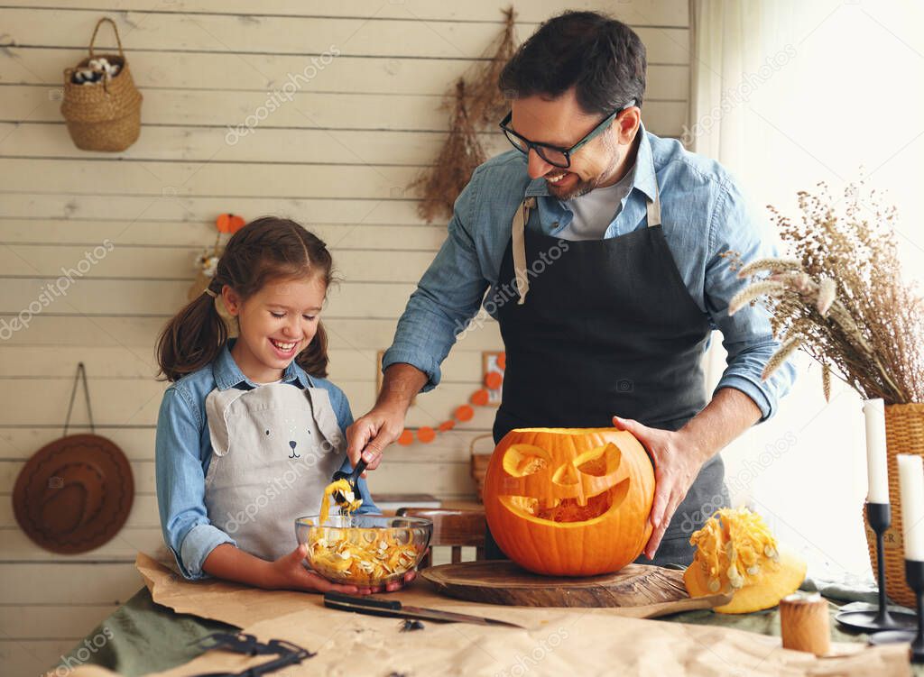Smiling father showing his little daughter how to carve pumpkin with spooky face, dad with child creating this iconic symbol of Halloween holiday jack-o-lantern while standing behind table in kitchen