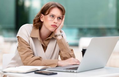 Kafeteryadaki terasta dizüstü bilgisayar ve defterle otururken internetten ders çalışan yorgun genç kız, kadın serbest çalışan yüz ifadesi olmadan yüzünü yan tarafa bakıyor.