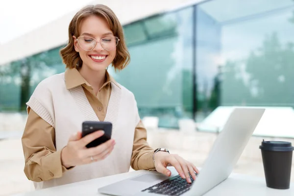Young happy business lady looks at smartphone screen with toothy smile while sitting outside on terrace with laptop and coffee, working remotely online checking reports and getting ready for meeting