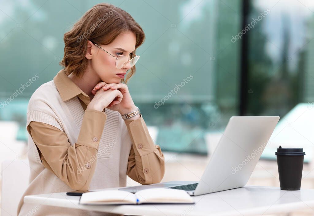 Serious concentrated young female office worker in glasses working remotely on laptop online sitting with takeaway coffee on terrace outdoors in cafe shop making notes from time to time in open agenda