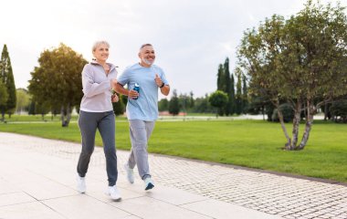 Full-length photo of lovely joyful retirees couple jogging outside in city park along alley with green trees, happy husband and wife looking at each other with smile holding water bottles in hands clipart