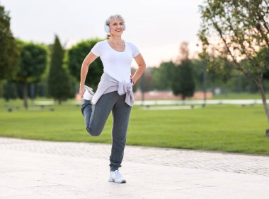 Kablosuz kulaklıklı olgun bir kadının fotoğrafı gülümseyerek bacak esnetme egzersizleri yapması, rutin antrenmandan önce kasları ısıtması ve güneşli bir sabahta şehir parkında koşması.