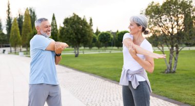 Sağlıklı zihin ve vücut. Güneşli bir sabahta parkta birlikte ısınırken spor giyim kuşamında gülümseyen olgun aile babası ve kadın kollarını esnetiyorlar. Etkin yaşam biçimi