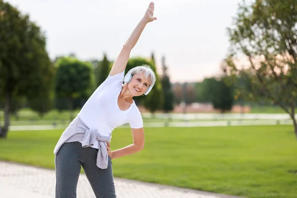 退休后继续保持活跃 快乐而成熟的退休女运动员 头戴耳机 身穿运动服 手挽手做侧身伸展运动 在公园外锻炼 快乐而又成熟 — 图库照片