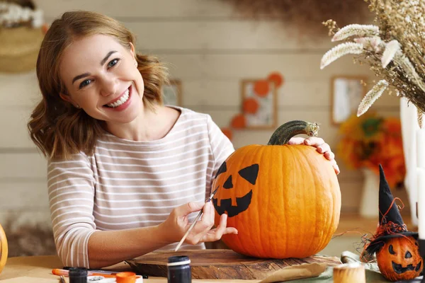 Young woman drawing scary face on orange pumpkin with paint brush for house decoration during Halloween holiday, female creating jack-o-lantern while sitting at table in kitchen at home
