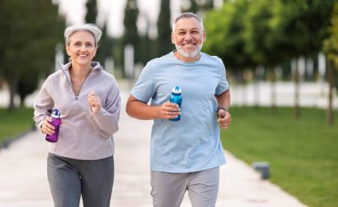 Lovely joyful retirees couple jogging outside in city park along alley with green trees, happy husband and wife looking at each other with smile holding water bottles in hands clipart
