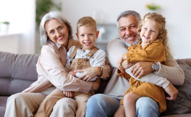 Mutlu aile, neşeli küçük çocuklar, büyük anne ve büyük babalarıyla kucaklaşırken evdeki oturma odasında birlikte oturuyorlar. Neşeli büyükanne ve büyükbaba kameraya gülümsüyorlar.