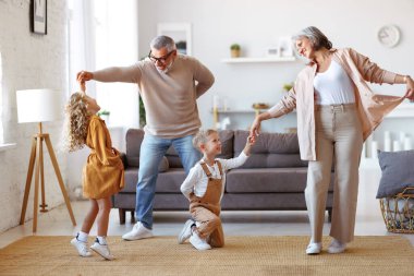 Aktif kıdemli büyükanne ve büyükbaba, oturma odasında iki mutlu torunla dans ediyor, küçük çocuklar, kız kardeş ve erkek kardeş oturma odasında büyük anne ve büyükbabayla oynarken eğleniyorlar.