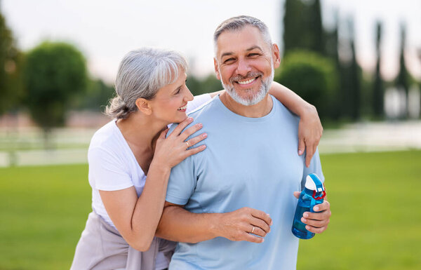Portrait of lovely happy elderly couple on morning run outside in city park, retirees wife and husband rejoice in active lifestyle, smiling woman tenderly embracing her spouse after routine jogging