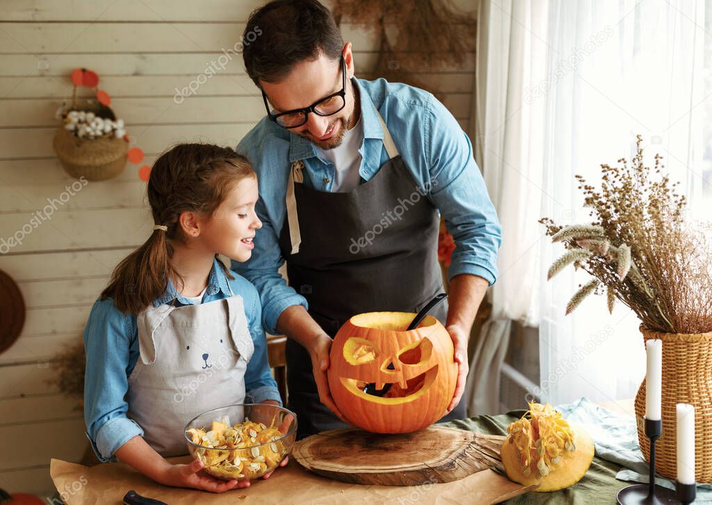 Smiling father showing his little daughter how to carve pumpkin with spooky face, dad with child creating this iconic symbol of Halloween holiday jack-o-lantern while standing behind table in kitchen