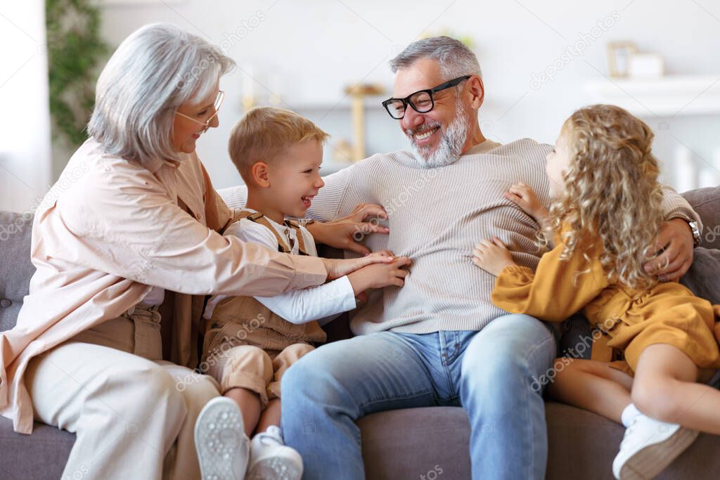 Playful kids and grandmother tickling grandpa while having fun at home, happy family senior grandparents and children having fun together and playing while relaxing resting on sofa in living room