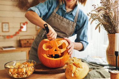 Cropped shot of woman in apron standing in kitchen and removing all the pulp from pumpkin with big spoon while carving jack o lantern in cozy kitchen at home, female making Halloween house decoration clipart