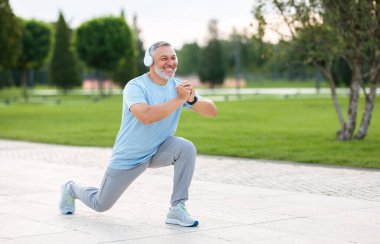Spor kıyafetli, kulaklıklı, mutlu, gülümseyen emekli bir sporcunun sabah rutin antrenman sırasında elleri bağlı bir şekilde şehir parkında tek ayak üstünde çömeldiği uzun bir fotoğraf.