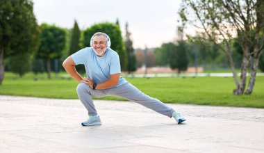 Açık hava antrenmanı sırasında mutlu, olgun sporcu, kulaklık takan, spor kıyafeti giyen, kaslarını ısıtan, tek bacağının üzerine çömelmiş, parkta aktif yaşam tarzının tadını çıkaran.