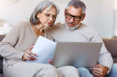 Ciddi bir aile çifti, evdeki kanepede otururken internet faturalarını ödüyor, emekli karı koca ellerinde mali belge tutuyor ve internet bankacılığını bilgisayar başında kullanıyor.