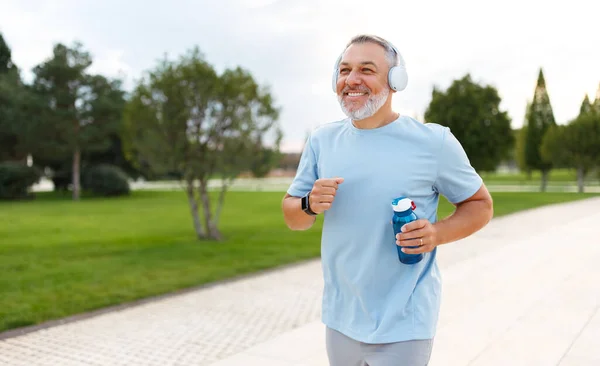 清晨在公园里慢跑时 戴着耳机的成熟男子快乐地拿着水瓶 面带微笑地跑着 享受着积极健康的生活方式 — 图库照片