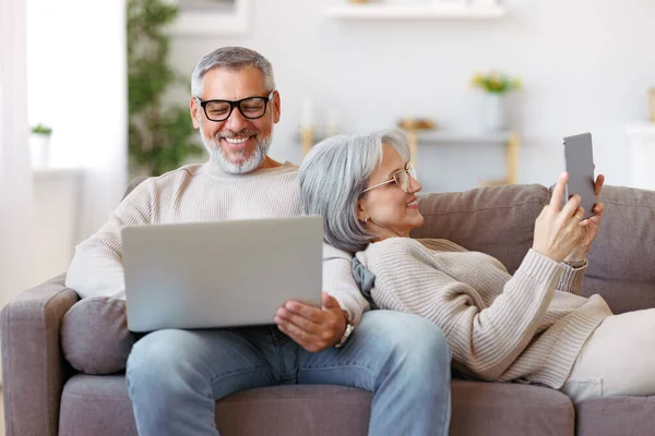 現代的な技術のラップトップとデジタルタブレットを使用して幸せなリラックスした古い退職した家族カップルリビングルームでソファでリラックスしながら 無料の時間でインターネットサーフィンシニア男性と女性 — ストック写真