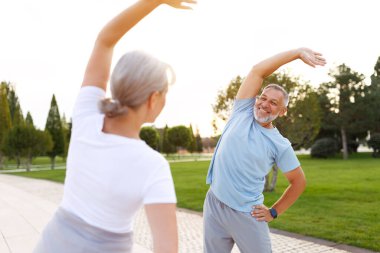 Sağlıklı zihin ve vücut. Güneşli bir sabahta parkta birlikte ısınırken spor giyim kuşamında gülümseyen olgun aile babası ve kadın kollarını esnetiyorlar. Etkin yaşam biçimi