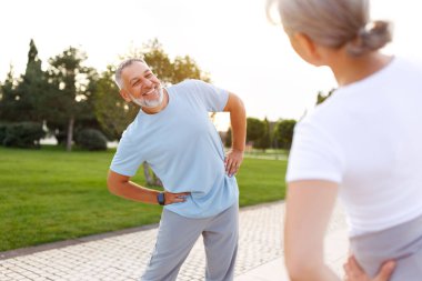 Sağlıklı zihin ve vücut. Güneşli bir sabahta parkta birlikte ısınırken spor giyim kuşamında gülümseyen olgun aile babası ve kadın kollarını esnetiyorlar. Etkin yaşam biçimi