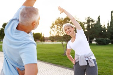 Sağlıklı zihin ve vücut. Güneşli bir sabahta parkta birlikte ısınırken spor giyim kuşamında gülümseyen olgun aile babası ve kadın kollarını esnetiyorlar. Etkin yaşam biçimi