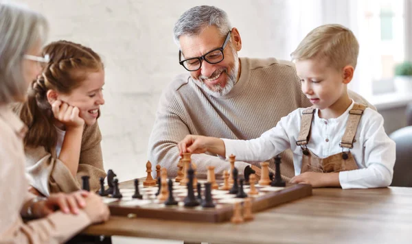 楽しい子供たち兄と妹は週末に一緒に時間を過ごしながら祖父母とリビングルームに座っている間にチェスをしています 子供たちはチェスボードと笑顔でテーブルに座っています — ストック写真