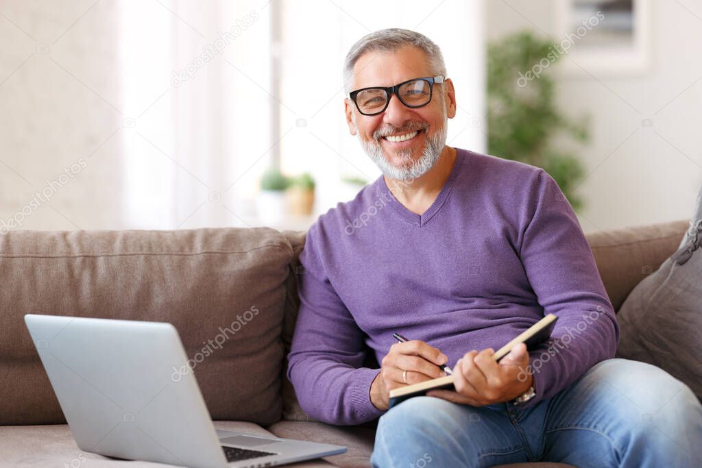 Handsome positive senior man wearing glasses working remotely while sitting on sofa with laptop computer at home, happy retired pensioner smiling at camera while learning studying online