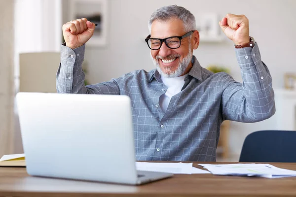 兴奋的老年人在庆祝成功 在远程教育期间取得了好的考试成绩 双手紧握拳头举手 坐在办公桌前 面带微笑地看着笔记本电脑 — 图库照片