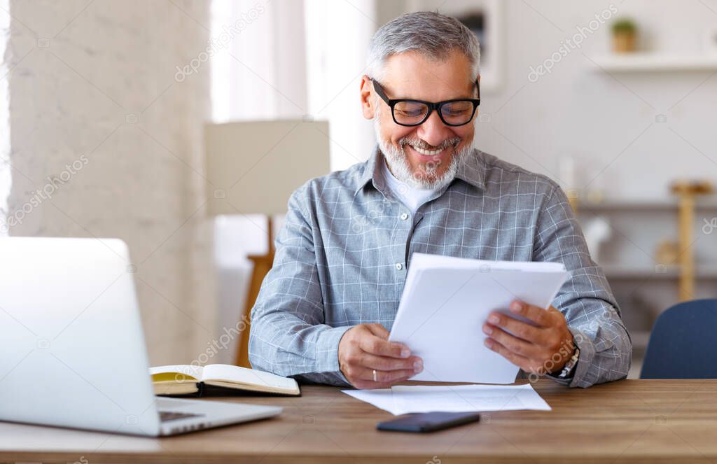 Handsome smiling senior man wearing glasses reading financial documents while working remotely on laptop at home, elderly businessman learning online, sitting at desk smile on face