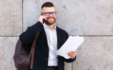 Deri çantalı, resmi kıyafetli genç işadamı müşterisiyle ya da ortağıyla cep telefonundan konuşuyor ve dışarıda beton duvarın önünde duran sözleşmeli belgeleri elinde tutuyor.