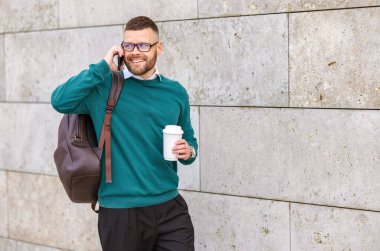Genç, mutlu bir ofis çalışanı cep telefonuyla konuşuyor elinde kahve, şehir binasının beton duvarına karşı duruyor, dışarıda akıllı telefonlu modern girişimci.