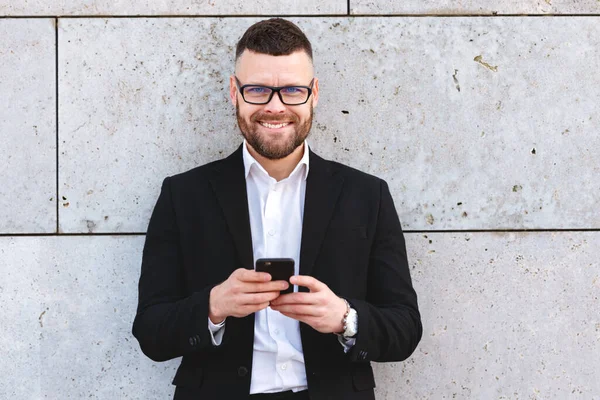 积极快乐的城市商业专业人士穿着时髦西装 站在混凝土墙边用智能手机 商人拿着手机用应用程序 输入电子邮件 — 图库照片