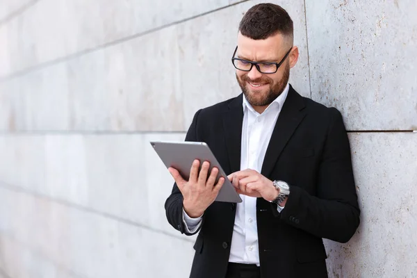 コンクリート壁の外に立っている間 デジタルタブレットを使用してスタイリッシュなスーツで積極的な陽気なビジネスマン 幸せなオフィスワーカーは 現代の電子デバイス上のメッセージメールを入力します — ストック写真